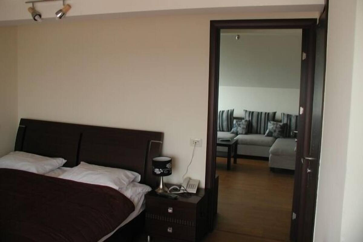 Un dormitorio con una cama grande, mesita de noche, lámpara y un espejo que refleja una sala de estar con sofá y almohadas.
