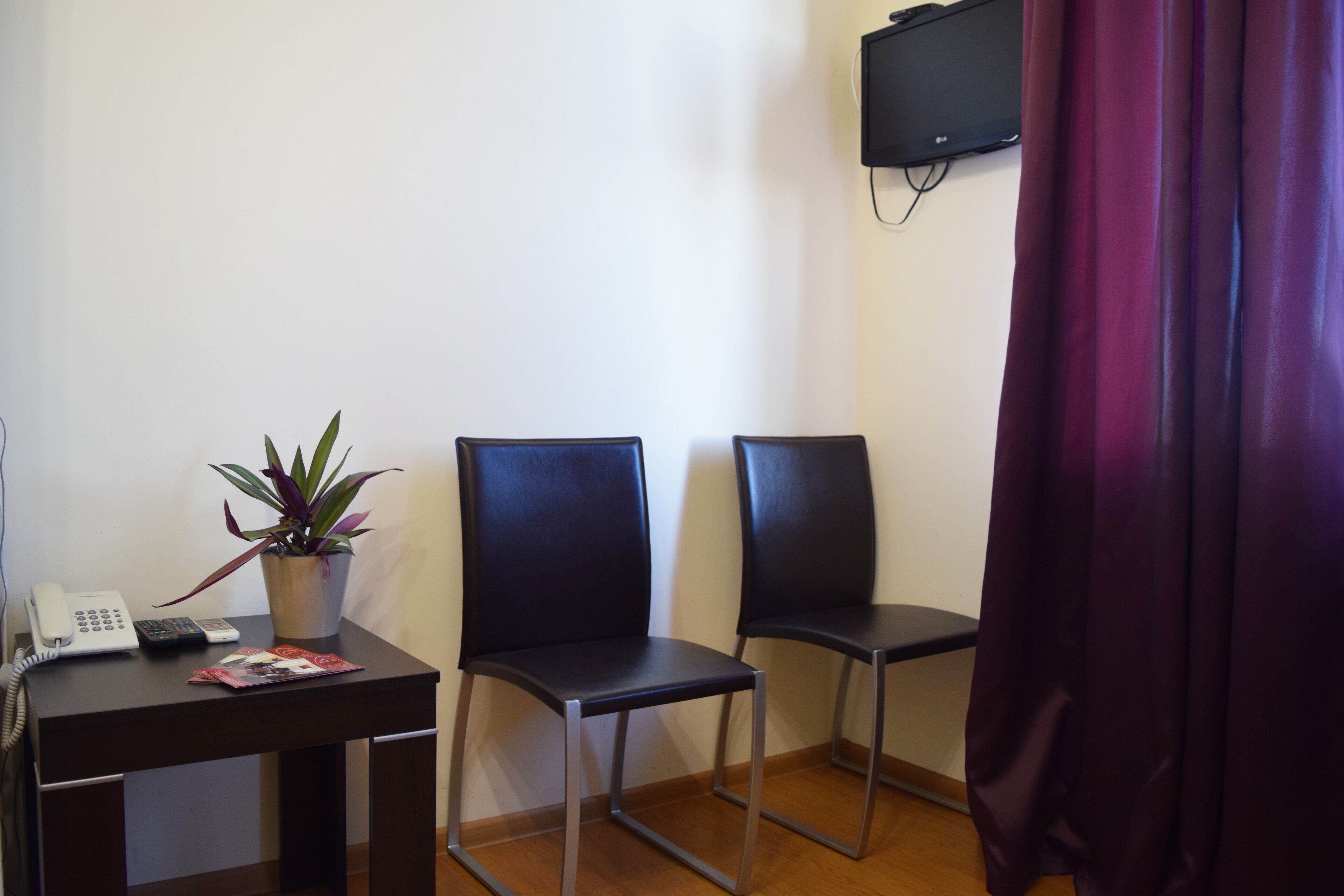 Una habitación con escritorio, dos sillas, una planta en maceta, teléfono y televisor montado en la pared.