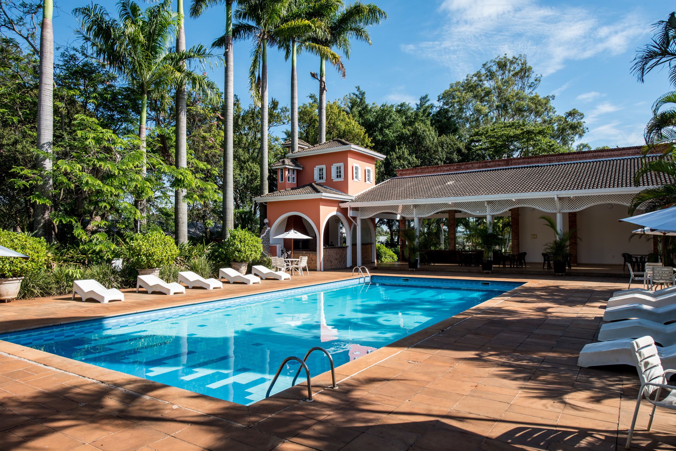 A swimming pool with lounge chairs, a covered patio, and a building with a tiled roof.