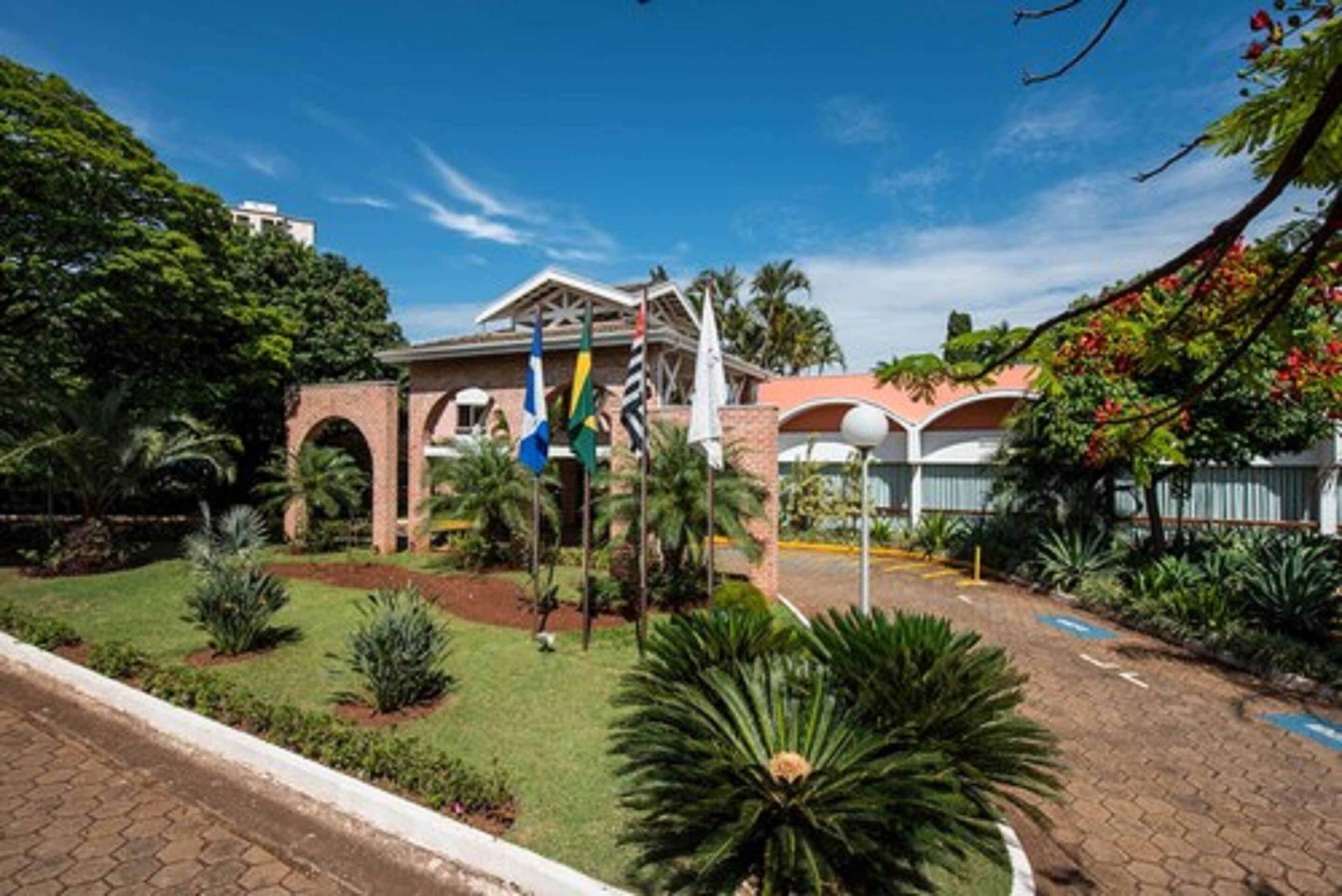 Un edificio de fachada de ladrillo, varias banderas y un jardín bien cuidado con diversas plantas.