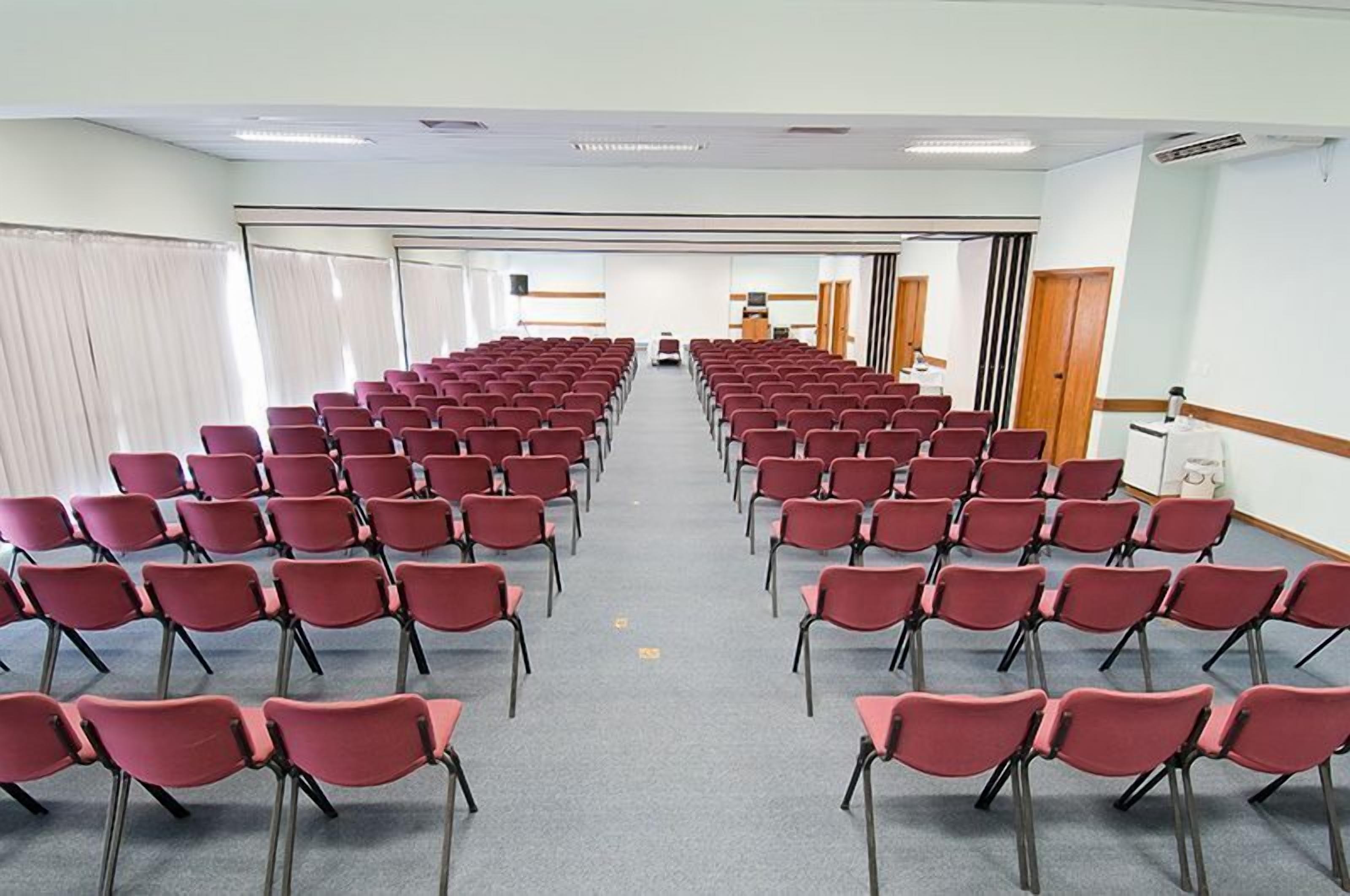 Un salón de conferencias con filas de sillas rojas, una pantalla de proyector y un atril al frente.