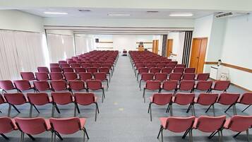 Un salón de conferencias con filas de sillas rojas, una pantalla de proyector y un atril al frente.
