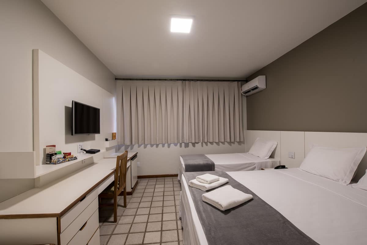 A hotel room with a desk, two beds, a TV, and an air conditioning unit.