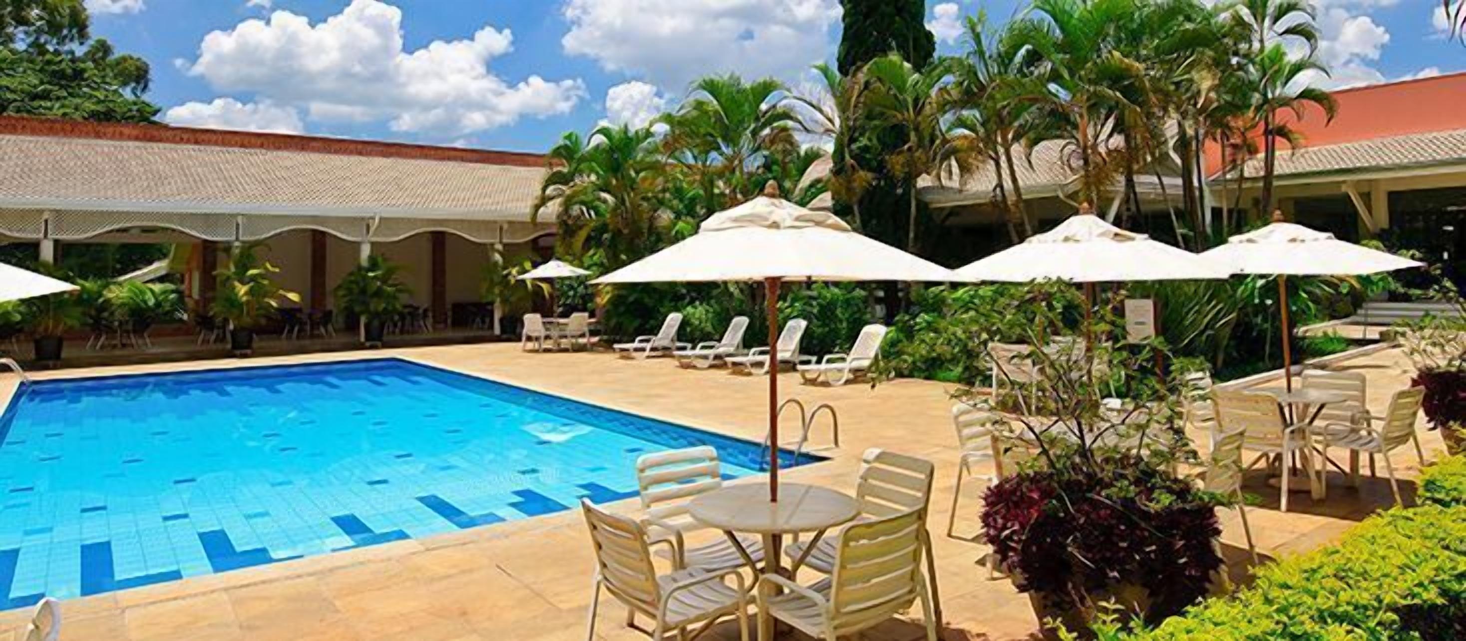 Una piscina con sillas de descanso y sombrillas, rodeada de palmeras y una zona cubierta.