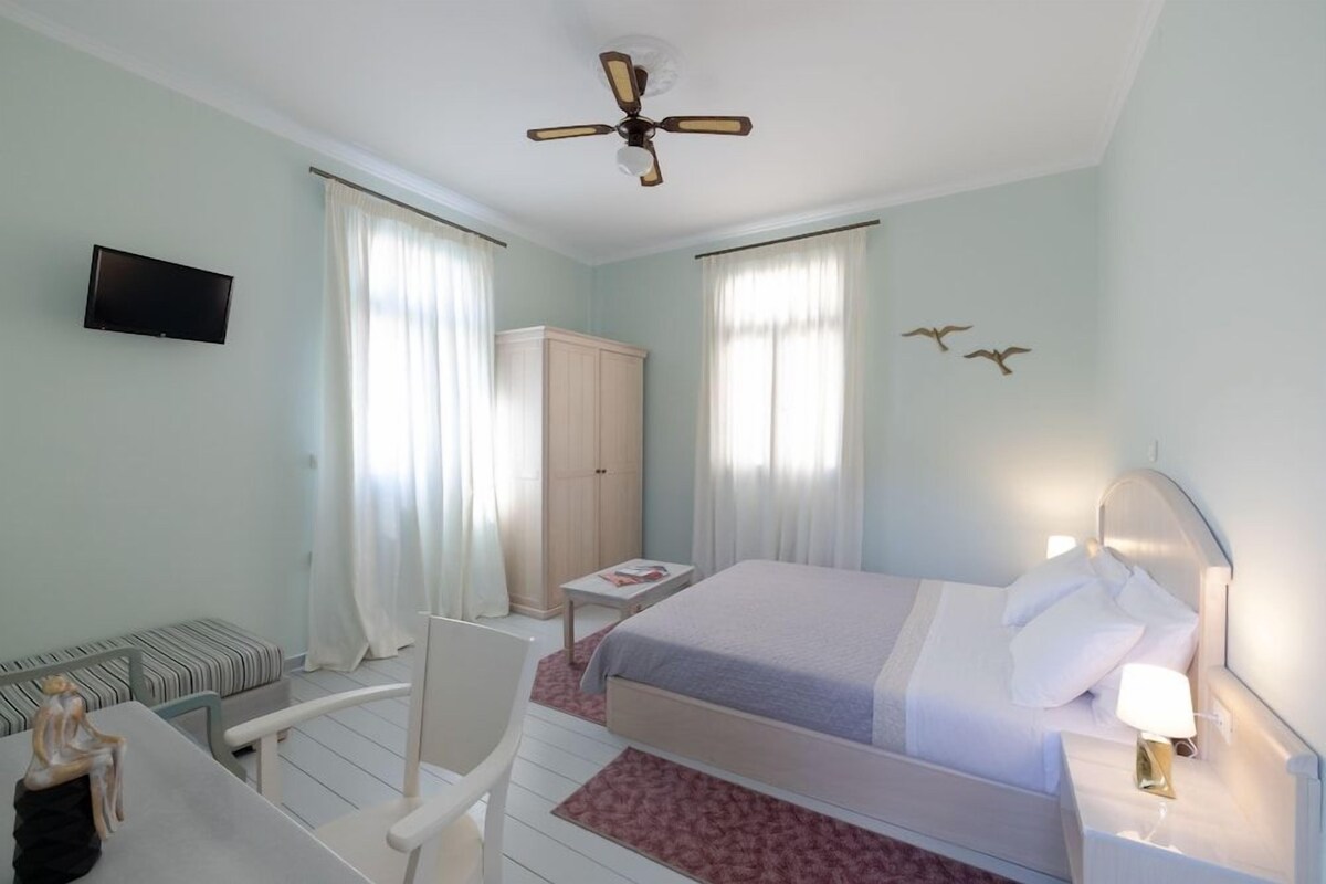 A hotel room with a bed, a desk, a chair, a television, a wardrobe, and a ceiling fan.