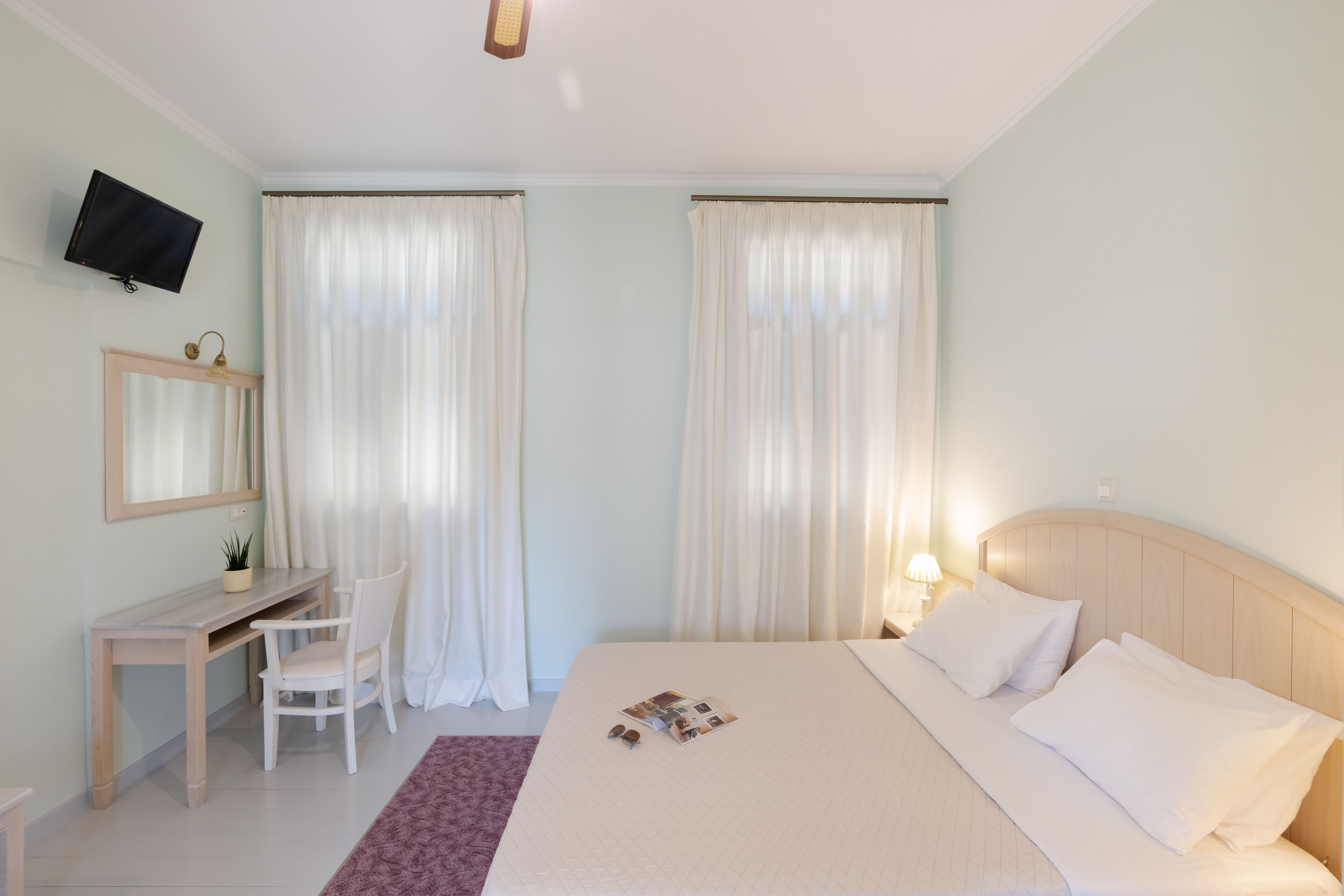 A hotel room with a bed, a desk with a chair, a television mounted on the wall, and curtains on the windows.