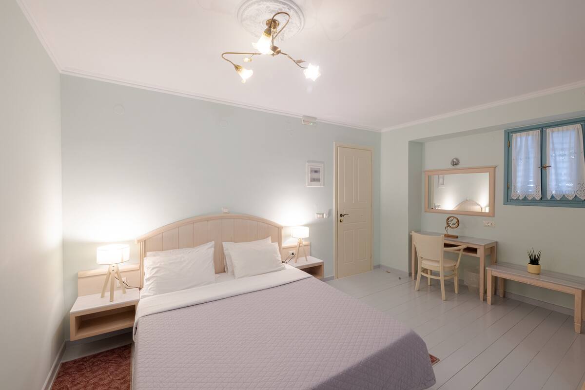 Un dormitorio con cama, mesitas de noche, escritorio, silla, espejo y lámpara en el techo.
