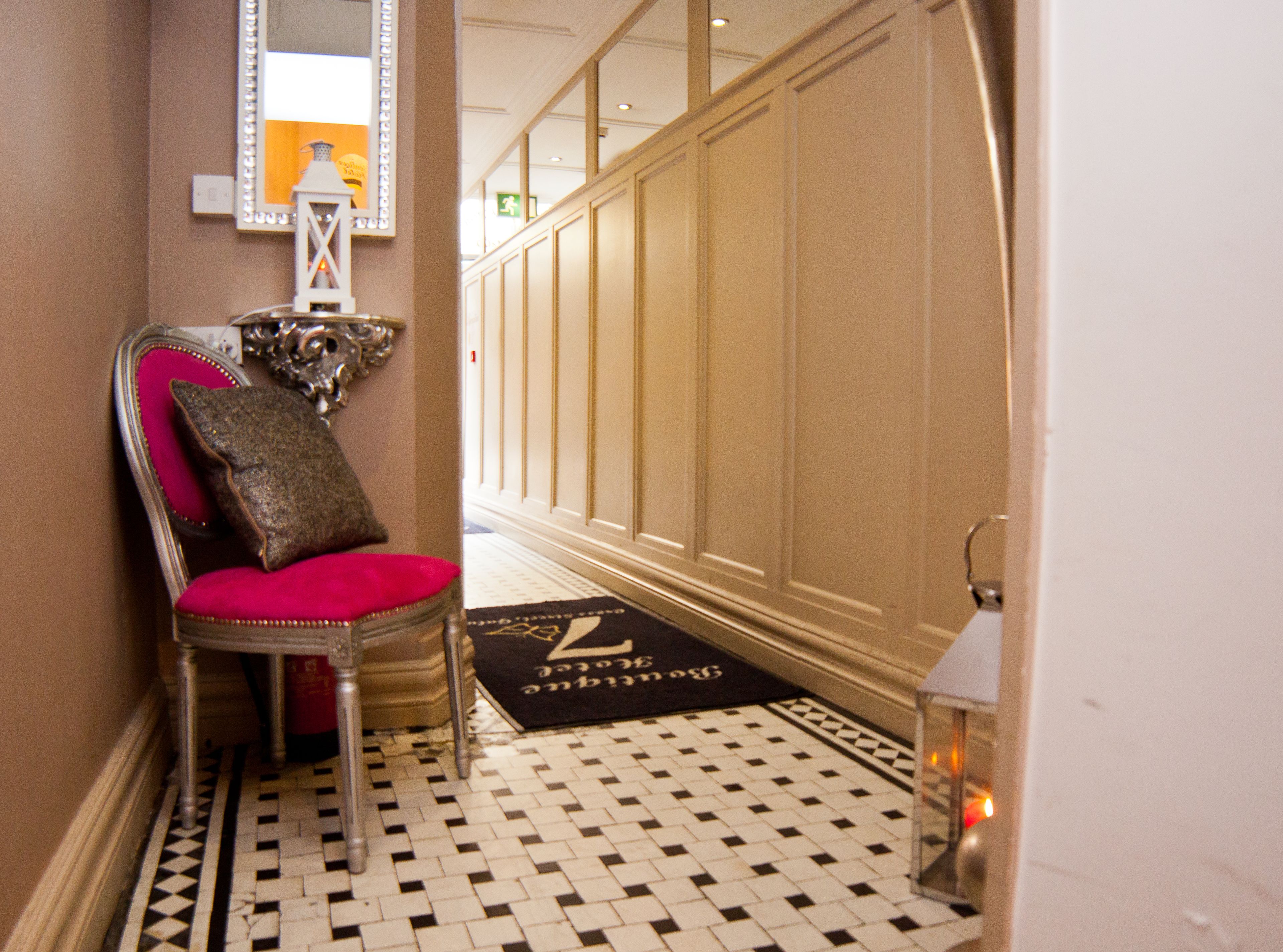 A hallway with a checkered floor, a red upholstered chair with a decorative pillow, a mirror, and a door with a doormat.