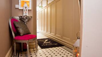 A hallway with a checkered floor, a red upholstered chair with a decorative pillow, a mirror, and a door with a doormat.
