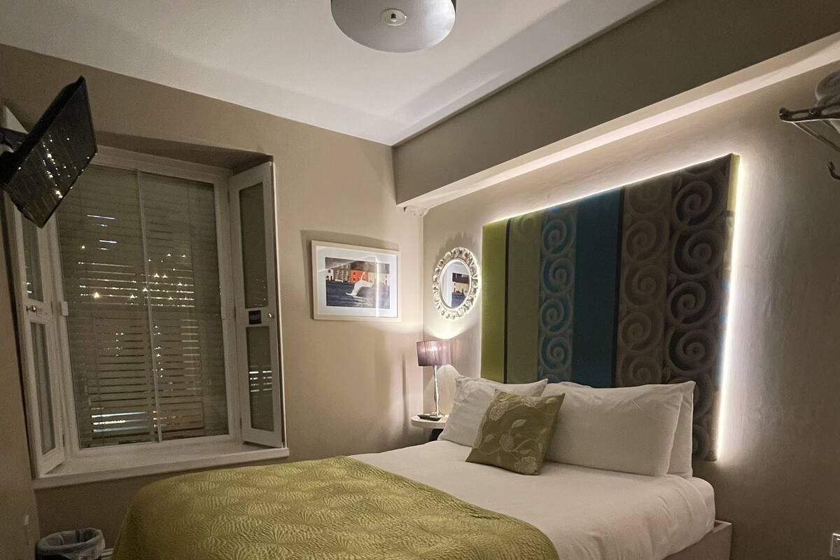 A bedroom with a bed, a bedside lamp, a window with blinds, a picture on the wall, and a ceiling light.