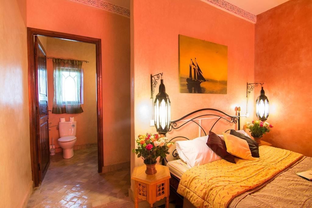 Une chambre à coucher avec un lit, une table de chevet avec une lampe, un tableau accroché au mur, et une salle de bain visible à travers une porte ouverte.