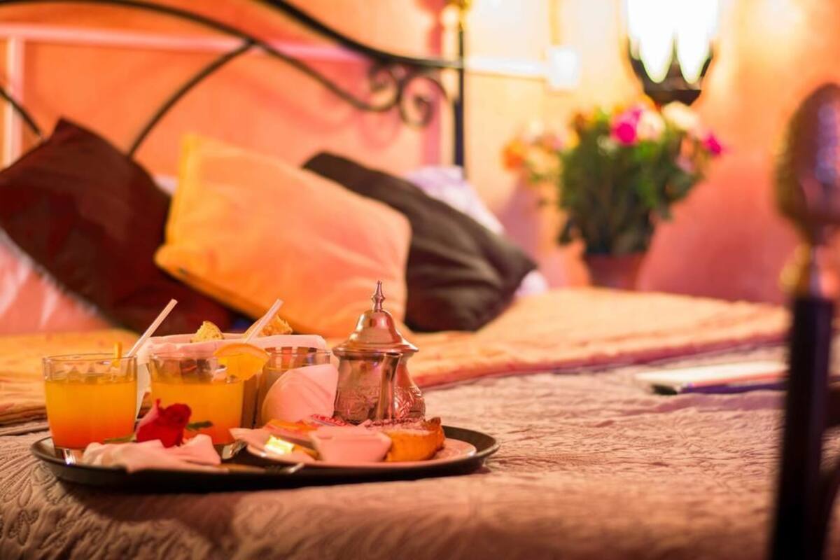 Una bandeja de desayuno con jugo de naranja, un pastel, y un azucarero sobre una cama con almohadas y una mesita de noche con una lámpara y flores.