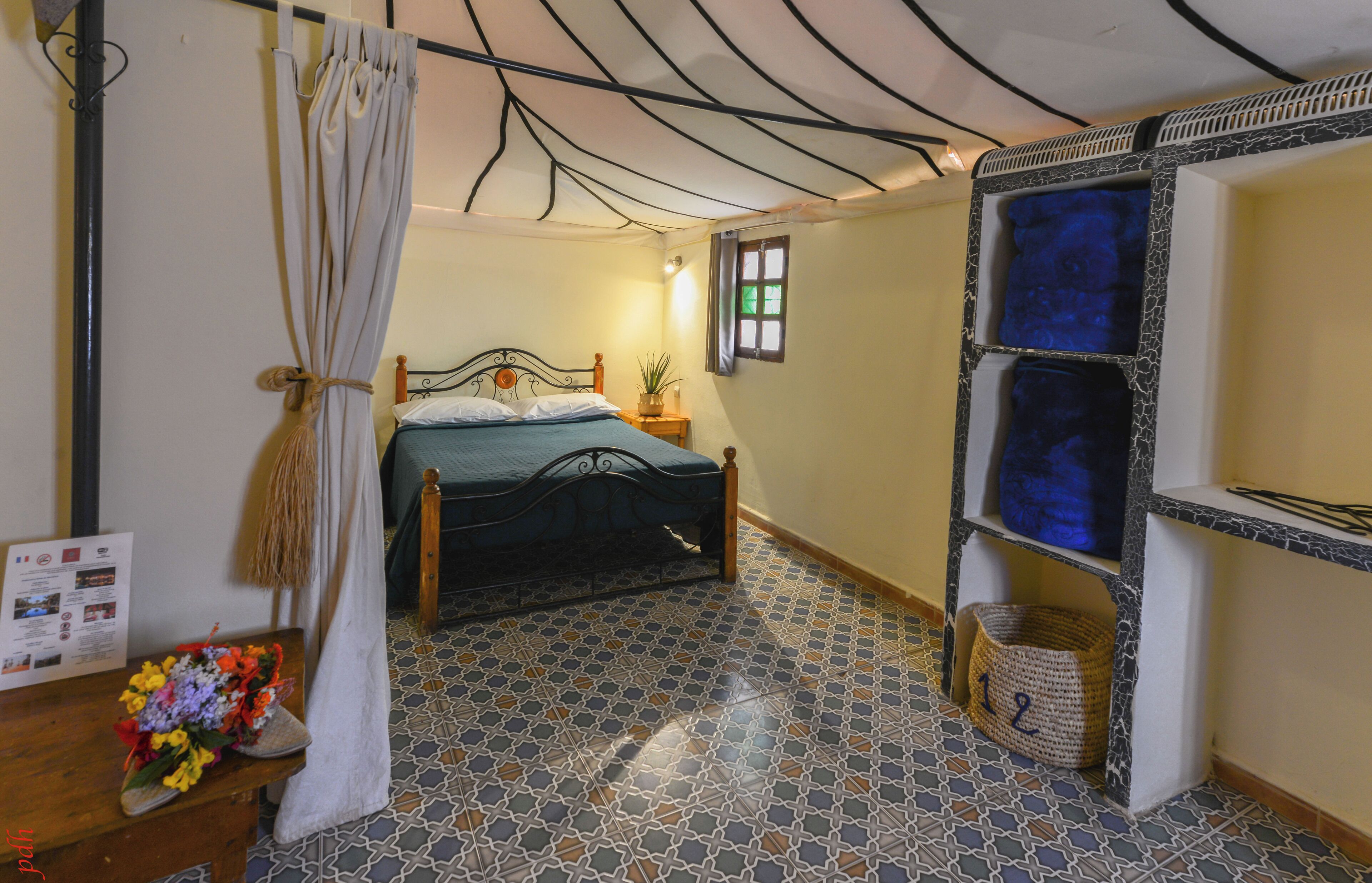 Een slaapkamer met een bed, nachtkastje, wandplank, een vloer met patroon en een gordijn.