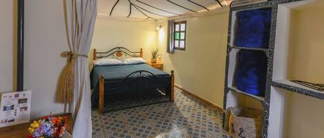 Een slaapkamer met een bed, nachtkastje, wandplank, een vloer met patroon en een gordijn.