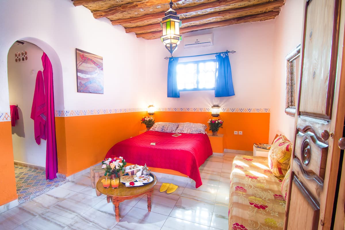 Un dormitorio con cama, una mesa con un plato de comida, una lámpara colgante y una ventana con cortinas.