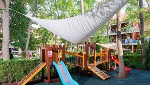 A playground with slides and climbing structures, surrounded by trees and a building in the background. - Zornica Residence (Sunny Beach)