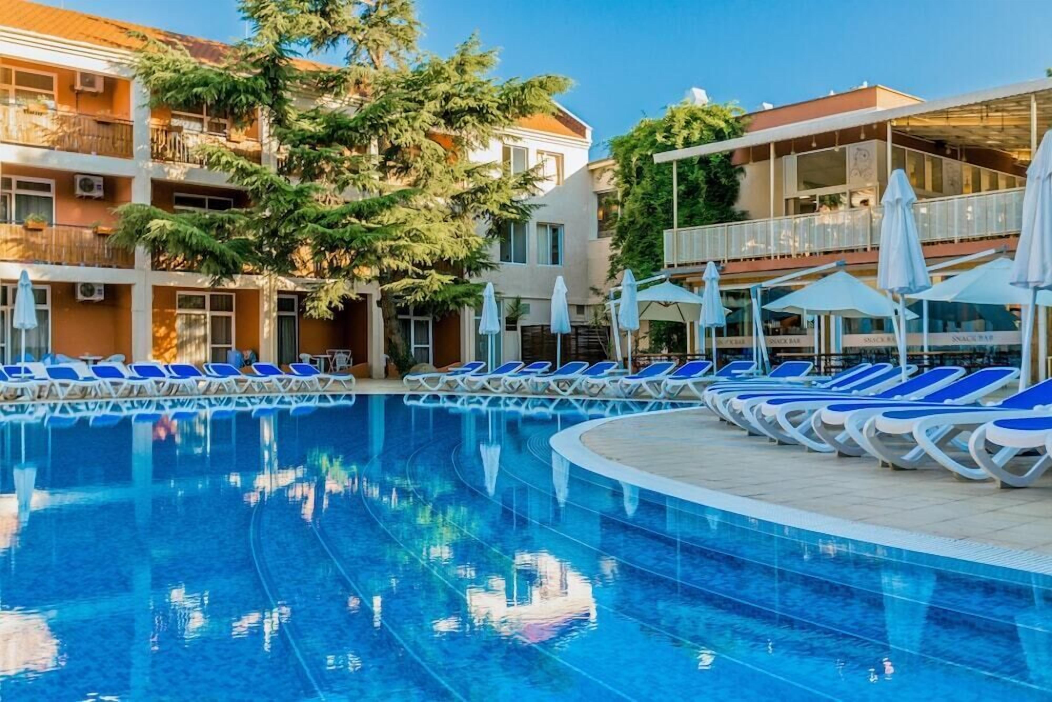 A swimming pool with lounge chairs and umbrellas, surrounded by multi-story buildings with balconies and outdoor seating areas.