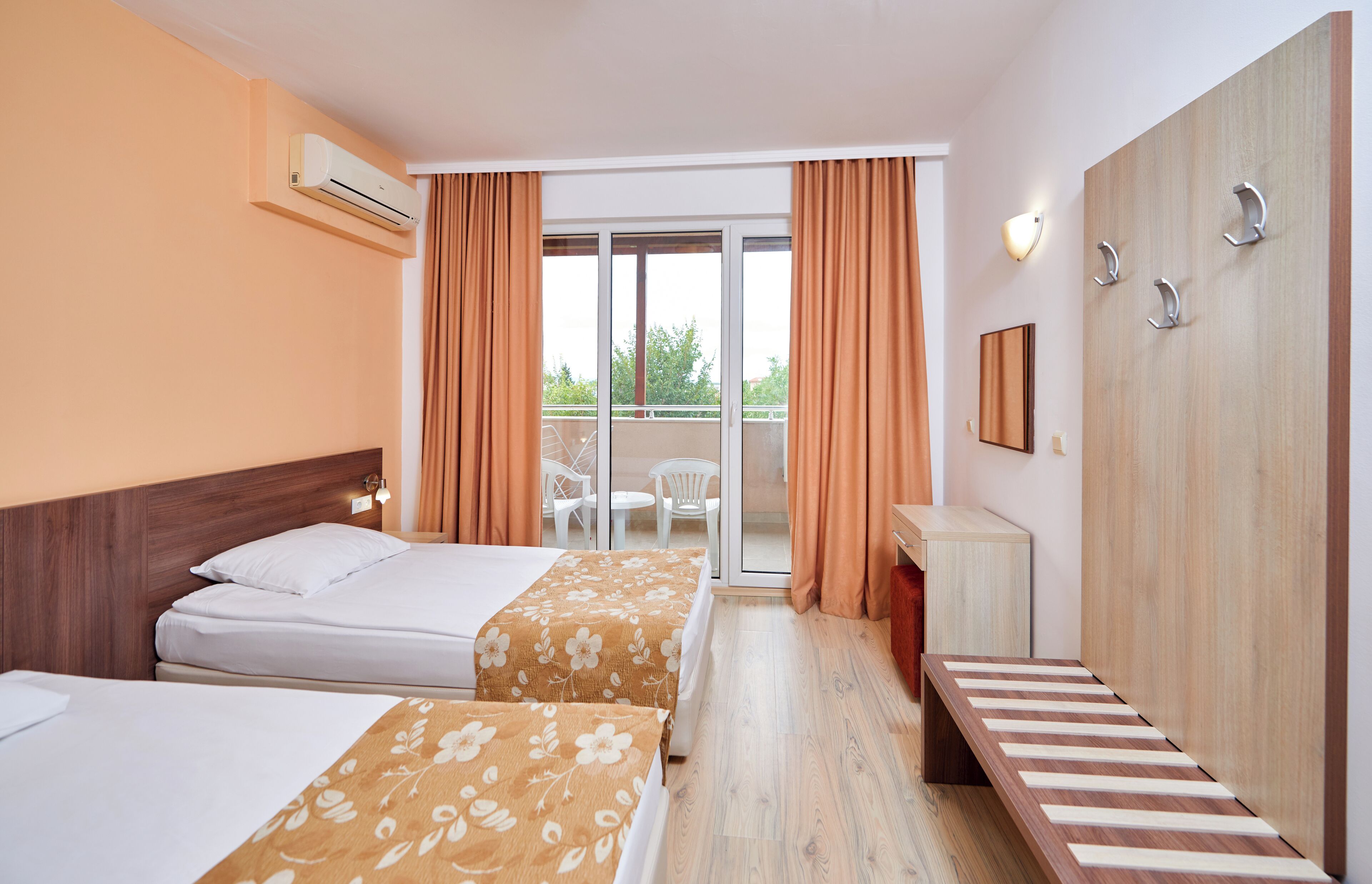 Hotellihuone, jossa on kaksi sänkyä, työpöytä, peili ja liukuovi, joka johtaa ulkotilaan, jossa on myös tuoleja.
