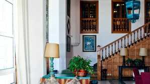 Un vaste espace de vie comprenant un ensemble de canapés, une table basse et un tapis à motifs. On y trouve un escalier avec des rampes en bois et un lustre.