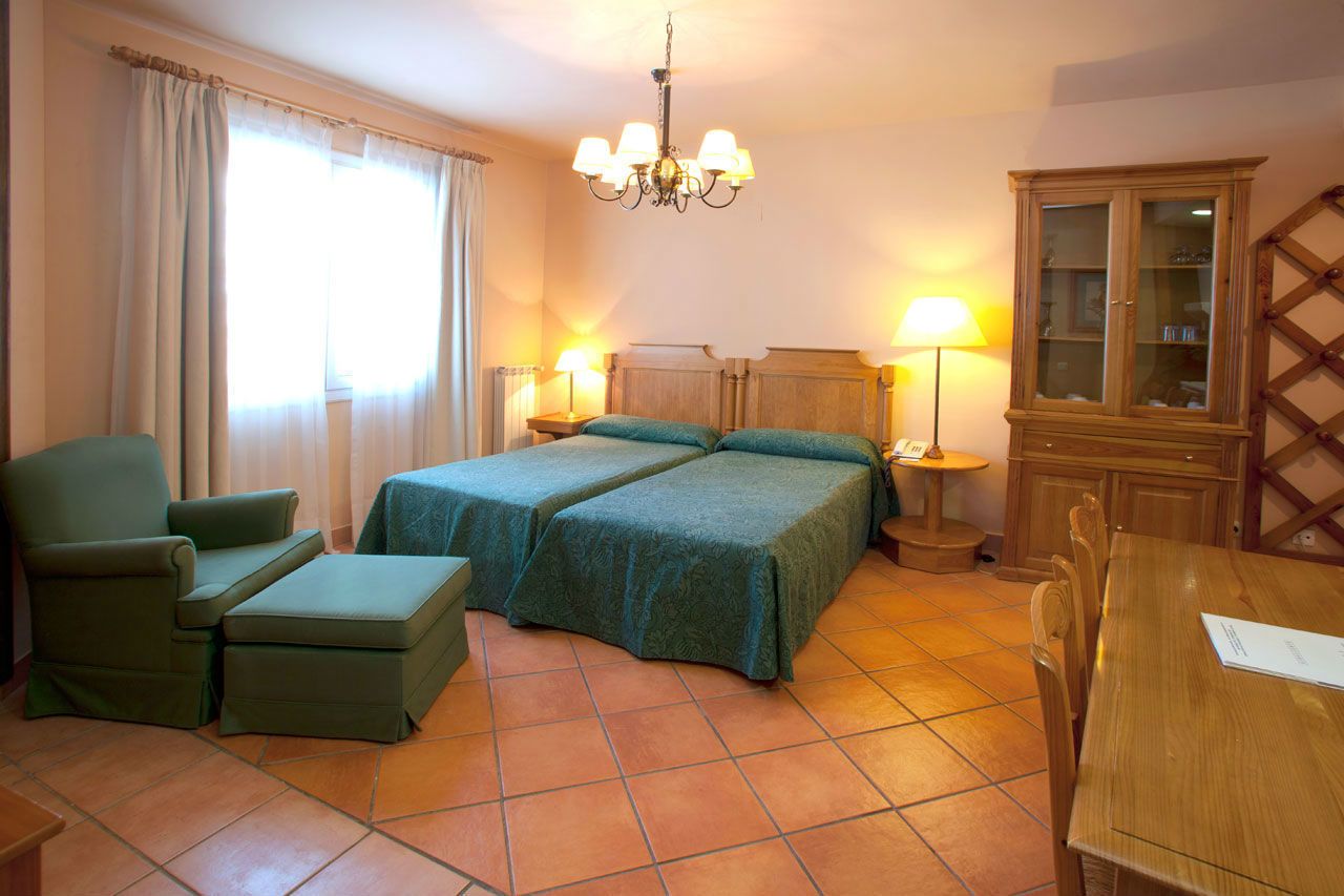 A hotel room with two beds, a green armchair, a wooden cabinet, and a dining table.