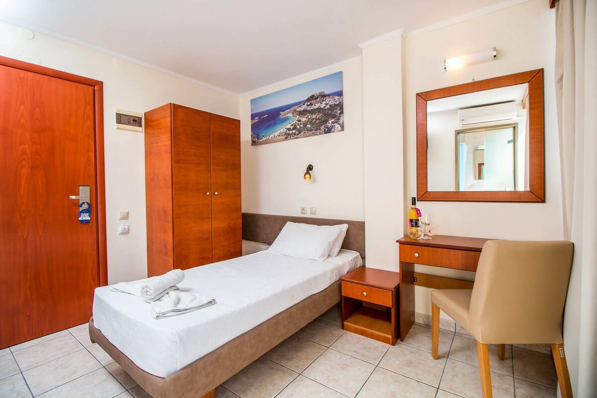 Habitación de hotel con una cama individual, armario de madera, escritorio con silla, espejo y un cuadro de un paisaje costero.