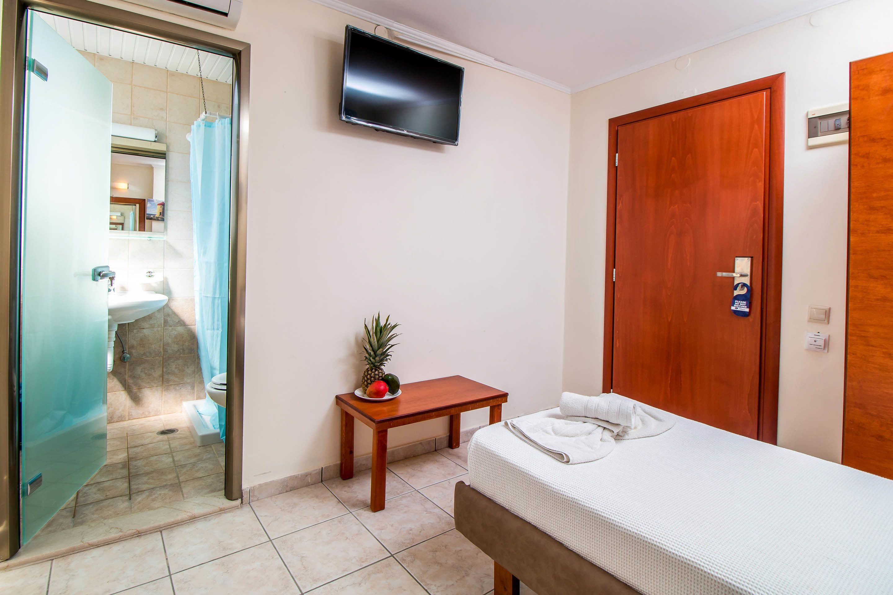 Camera d'albergo con un letto, una porta in legno, una TV a muro, un piccolo tavolo di legno con della frutta e un bagno visibile attraverso una porta aperta.