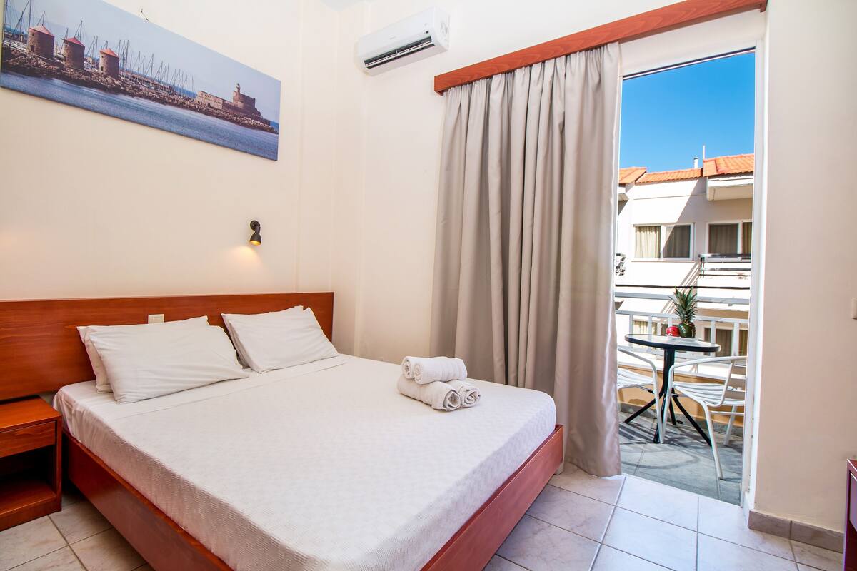 Habitación de hotel con cama, mesita de noche, un cuadro de una escena costera, aire acondicionado y balcón con mesa y sillas.