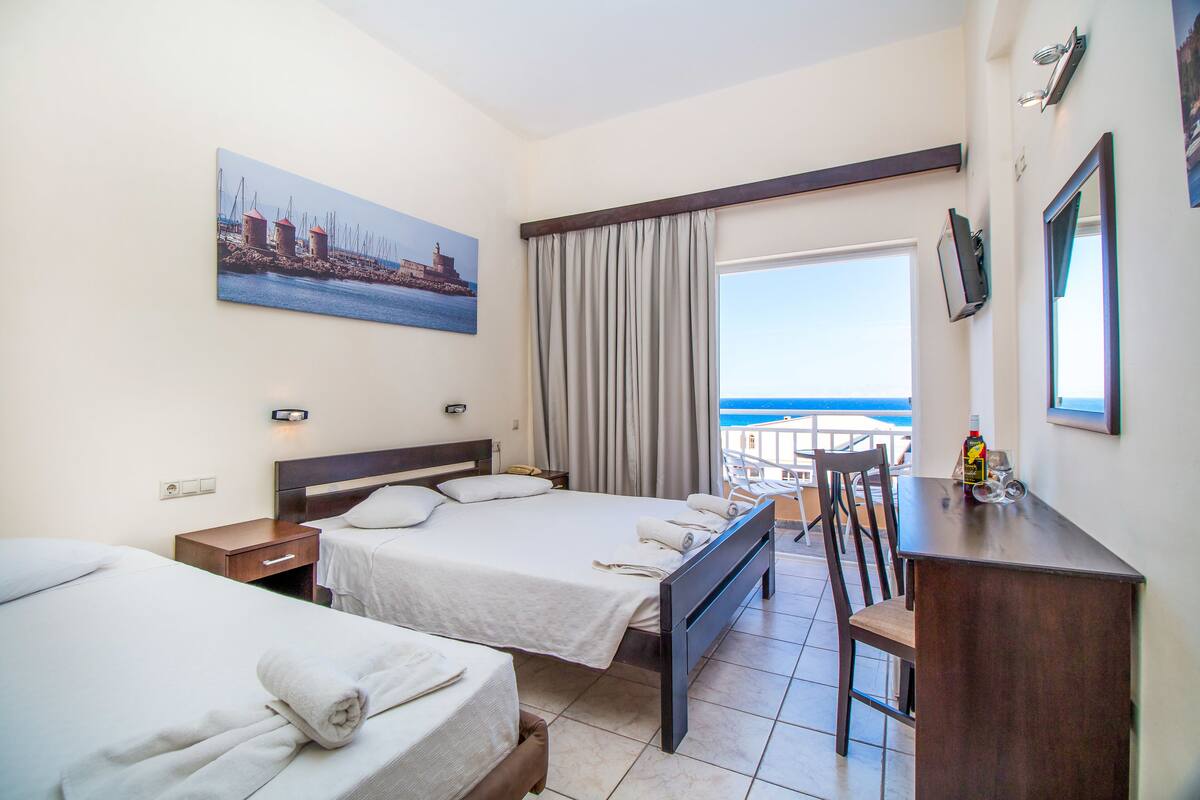 Habitación de hotel con dos camas, un escritorio, una silla, un televisor y una ventana con vista al mar.