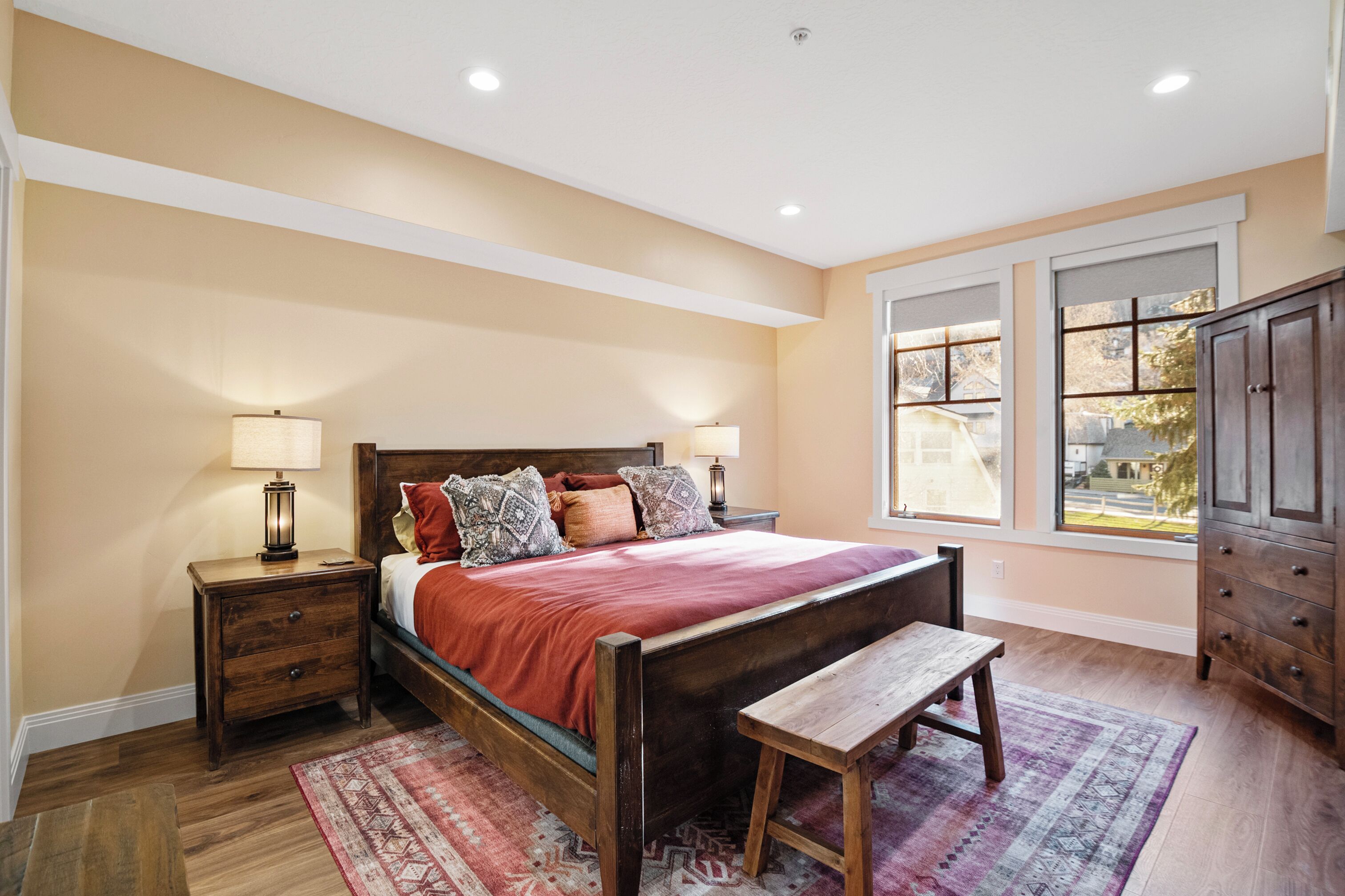 A bedroom with a wooden bed, two bedside tables with lamps, a bench, a large window, and a view of the outside.