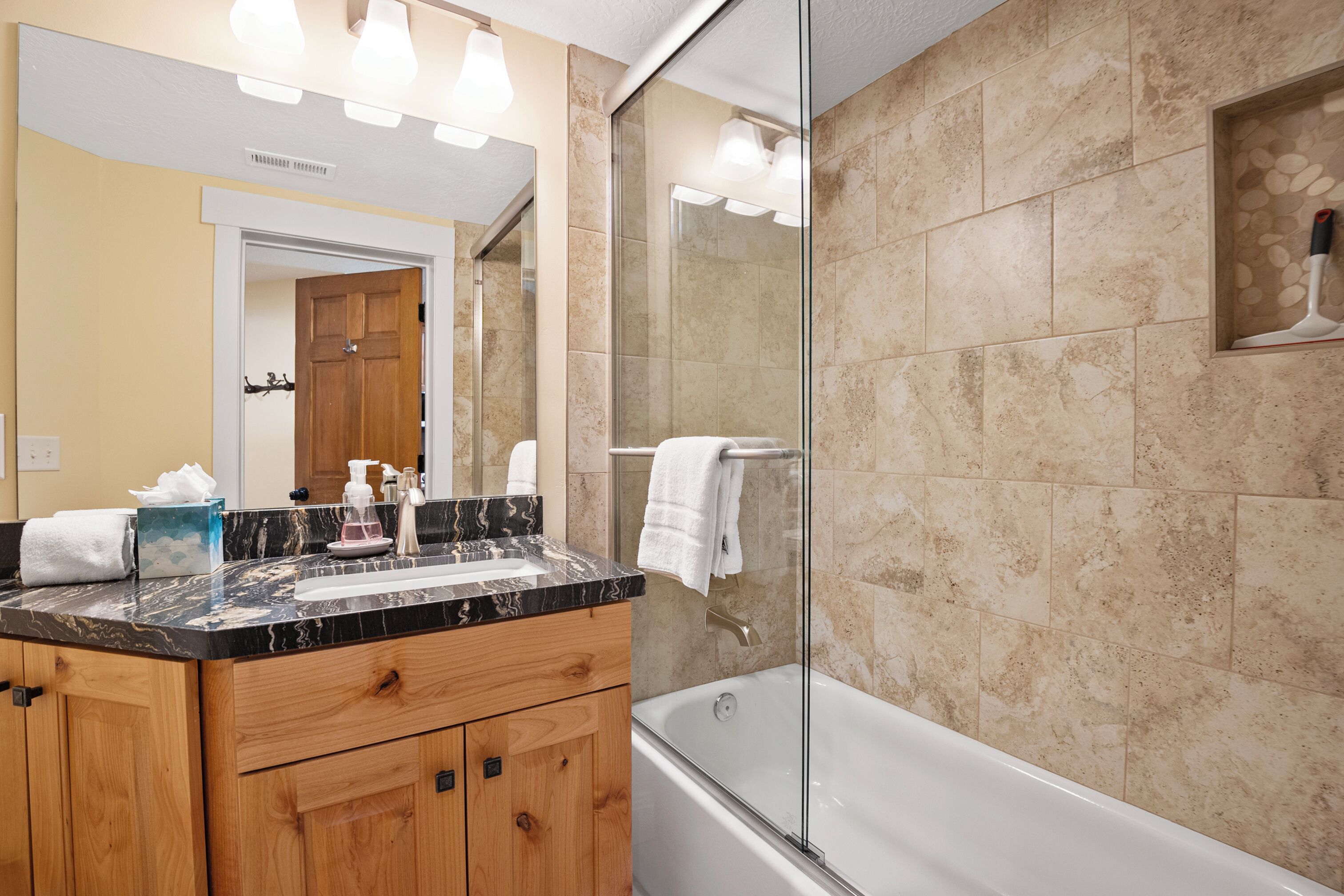 A bathroom with a marble countertop, wooden cabinets, a glass-enclosed shower, and beige tiled walls.