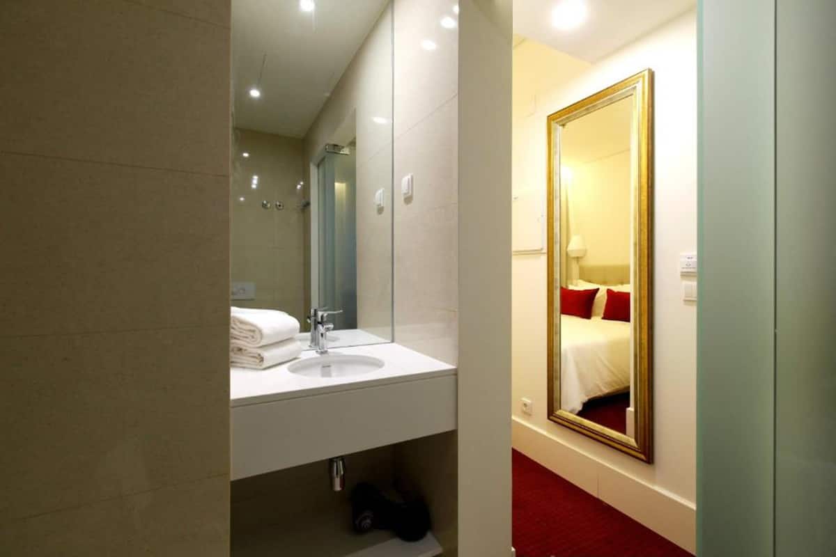 A hotel bathroom with a sink, mirror, and a large mirror reflecting a bed with red pillows.