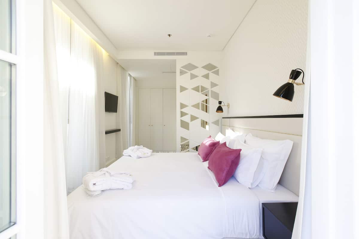 A hotel room with a bed, white linens, a pink throw, a black lamp, and a geometric wall design.