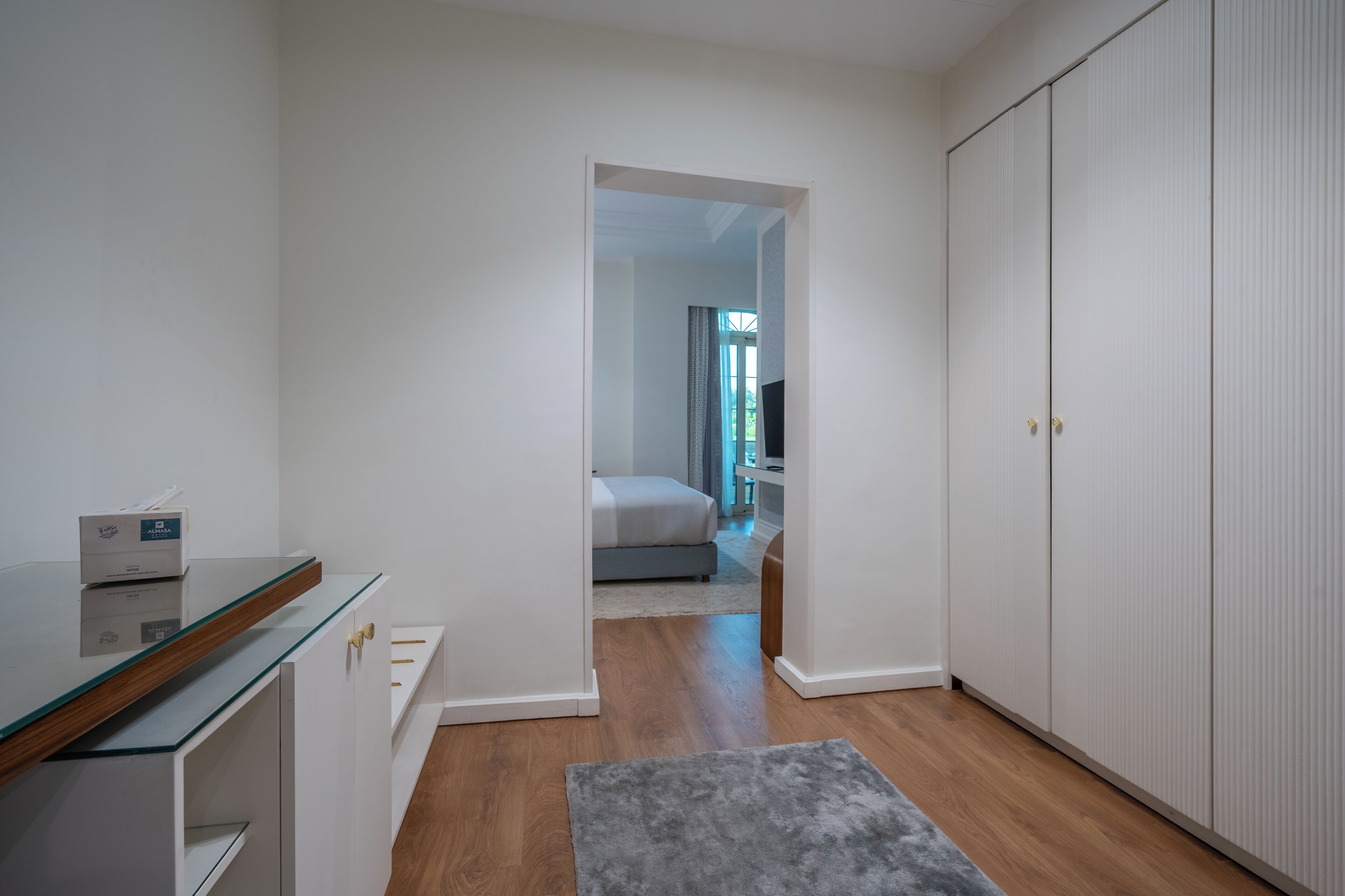 A hotel room with a glass-top desk, a wardrobe, and a bed in the background.