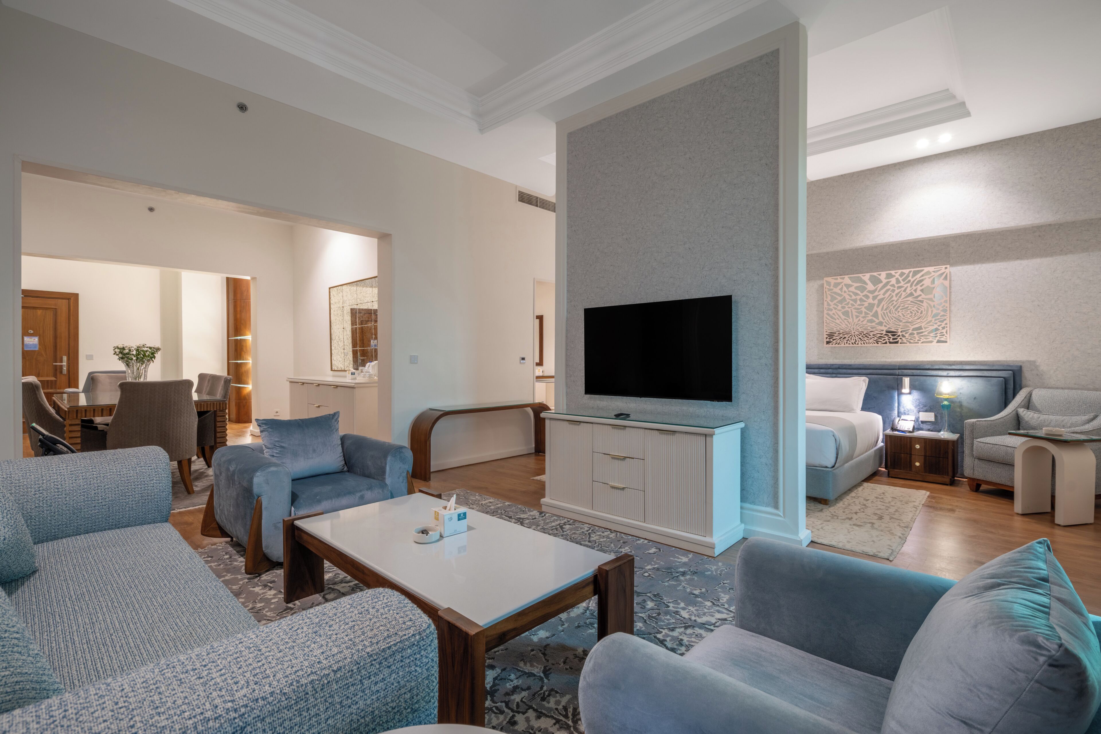 A modern living room with a flat-screen TV, a dining area with a table and chairs, and a separate bedroom with a bed and nightstand.