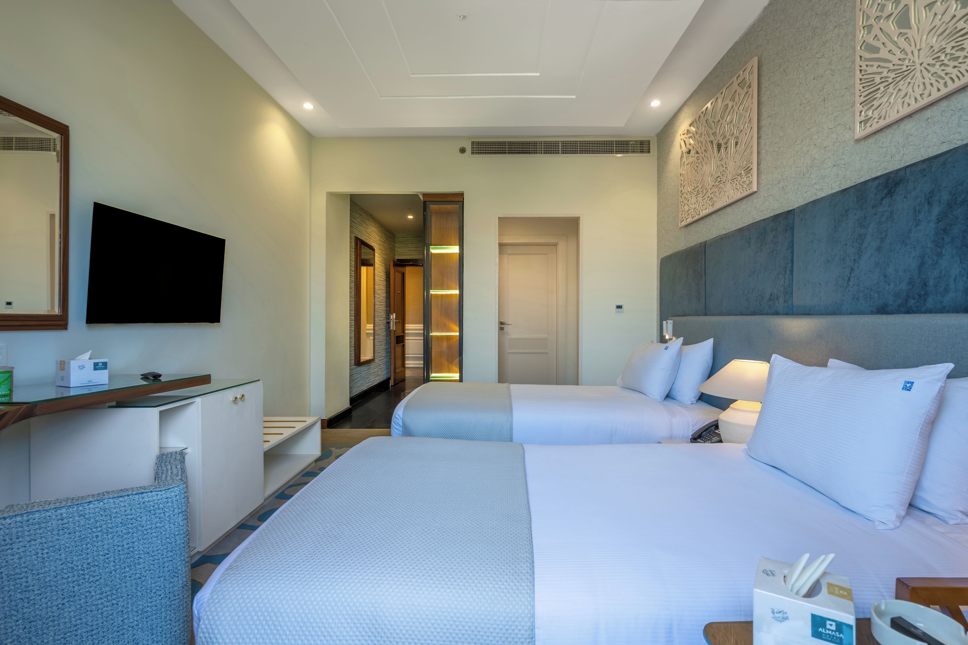 Une chambre d’hôtel avec deux lits, un bureau doté d’un téléviseur à écran plat, un miroir et un panneau décoratif fixé au mur.