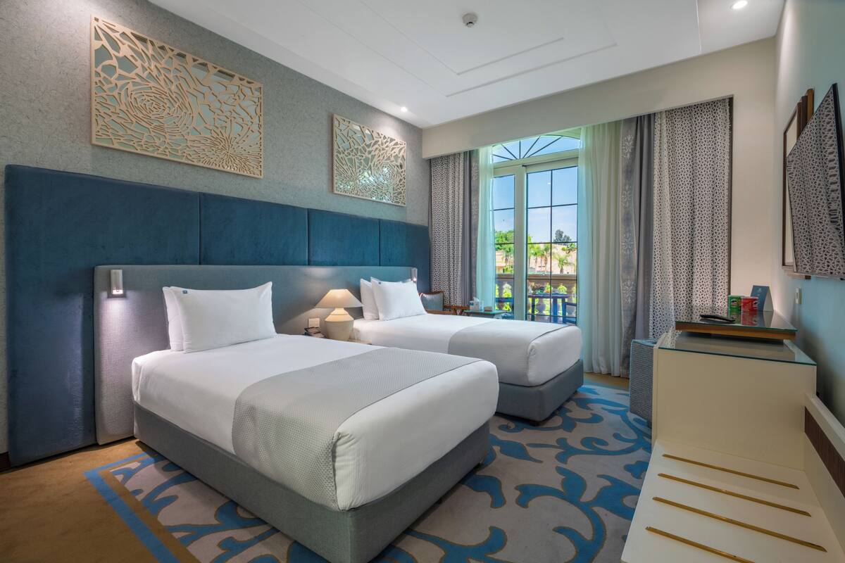 Une chambre d’hôtel avec deux lits, un bureau et une grande fenêtre avec des rideaux.