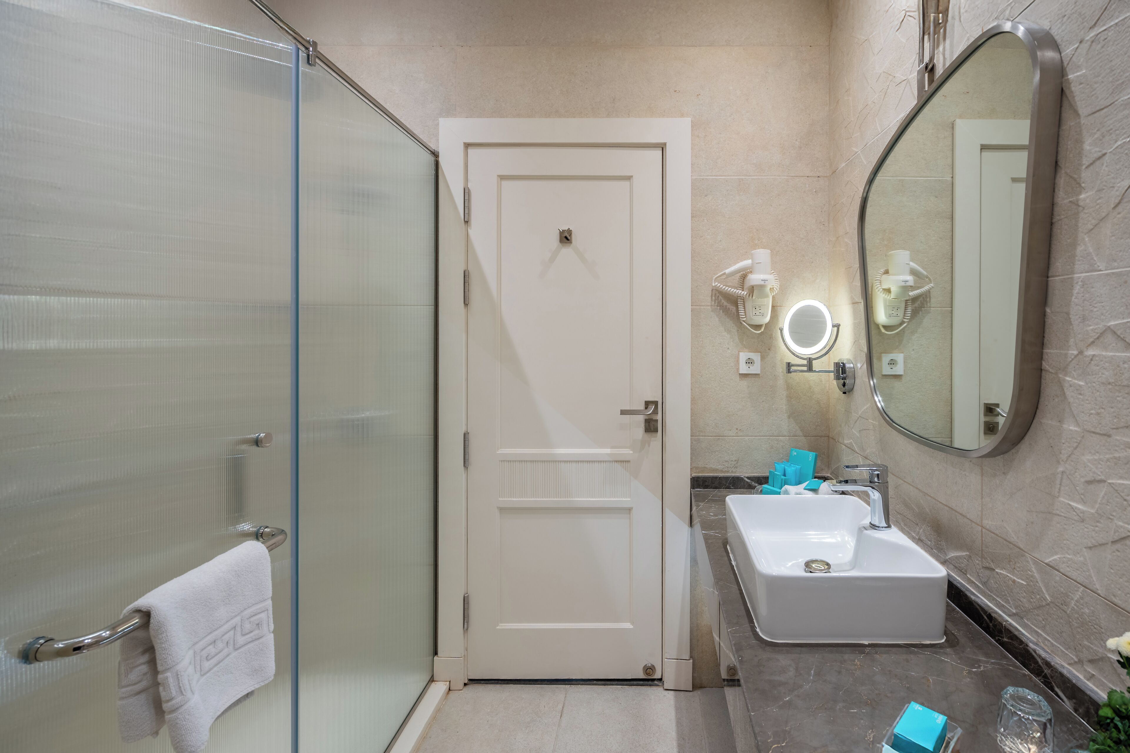 A bathroom with a glass shower enclosure, a white door, a sink with a mirror, and a towel rack.