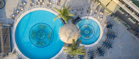 Dos piscinas grandes y circulares con una fuente central, rodeadas de zonas de estar al aire libre y palmeras.