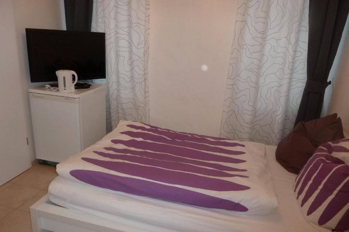 A neatly made bed with a white and purple striped bedspread, a small bedside table with a white kettle, and a television on a white cabinet.
