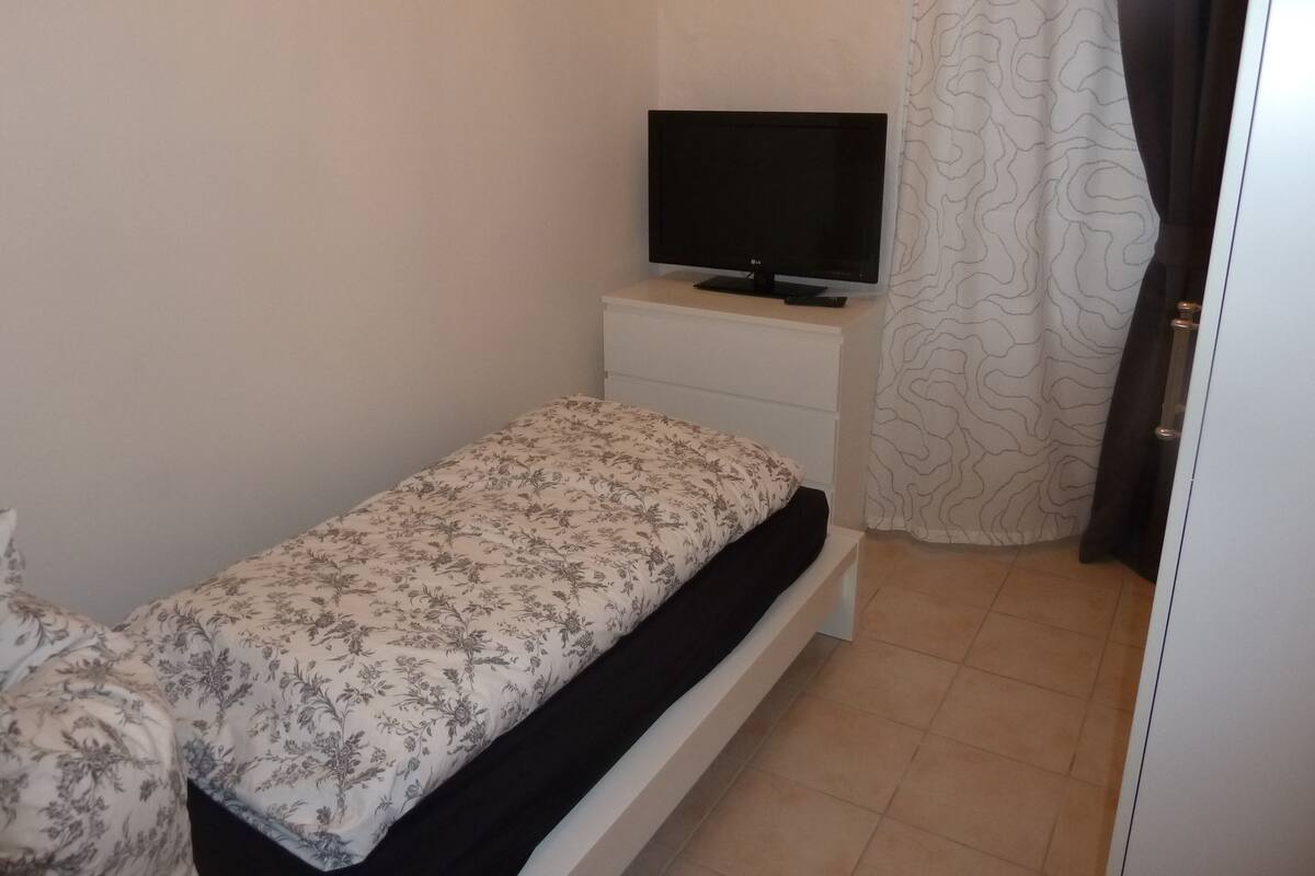A small bedroom with a single bed, a TV on a white nightstand, and a patterned curtain.
