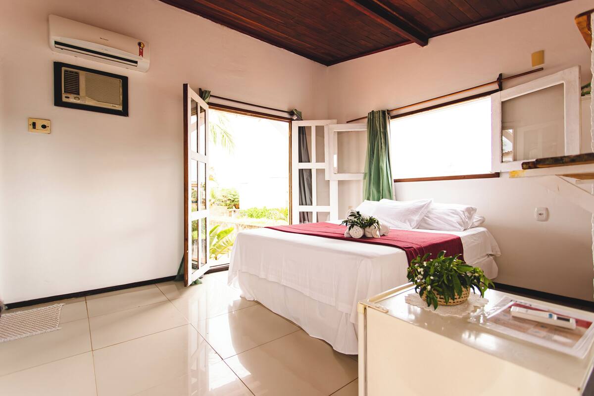 Un dormitorio con cama, mesita de noche con planta, ventana con cortinas y equipo de aire acondicionado.