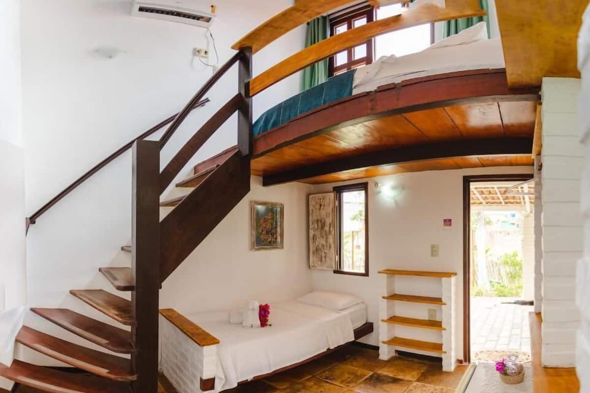 Una habitación con una escalera de madera, una cama, una mesita y un ventanal.