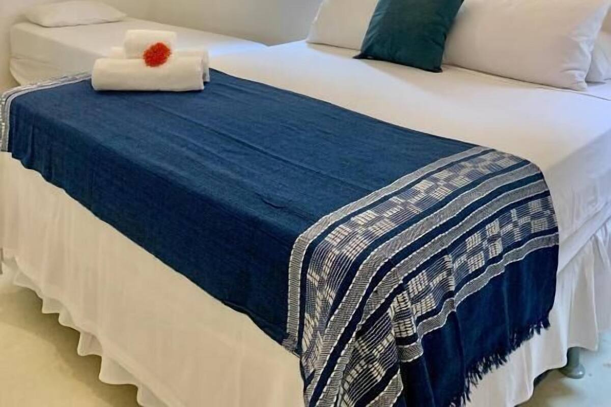 Un dormitorio con cama, almohadas, una manta azul y blanca estampada, una pared blanca, una ventana y un aire acondicionado instalado en la pared.