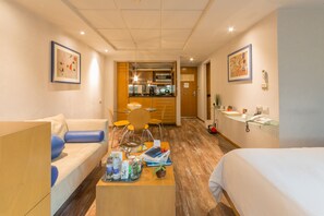 A hotel room with a sofa, a coffee table with drinks and magazines, a bed, a desk with a phone, and a kitchenette in the background. - Suites Contempo (Mexico City)