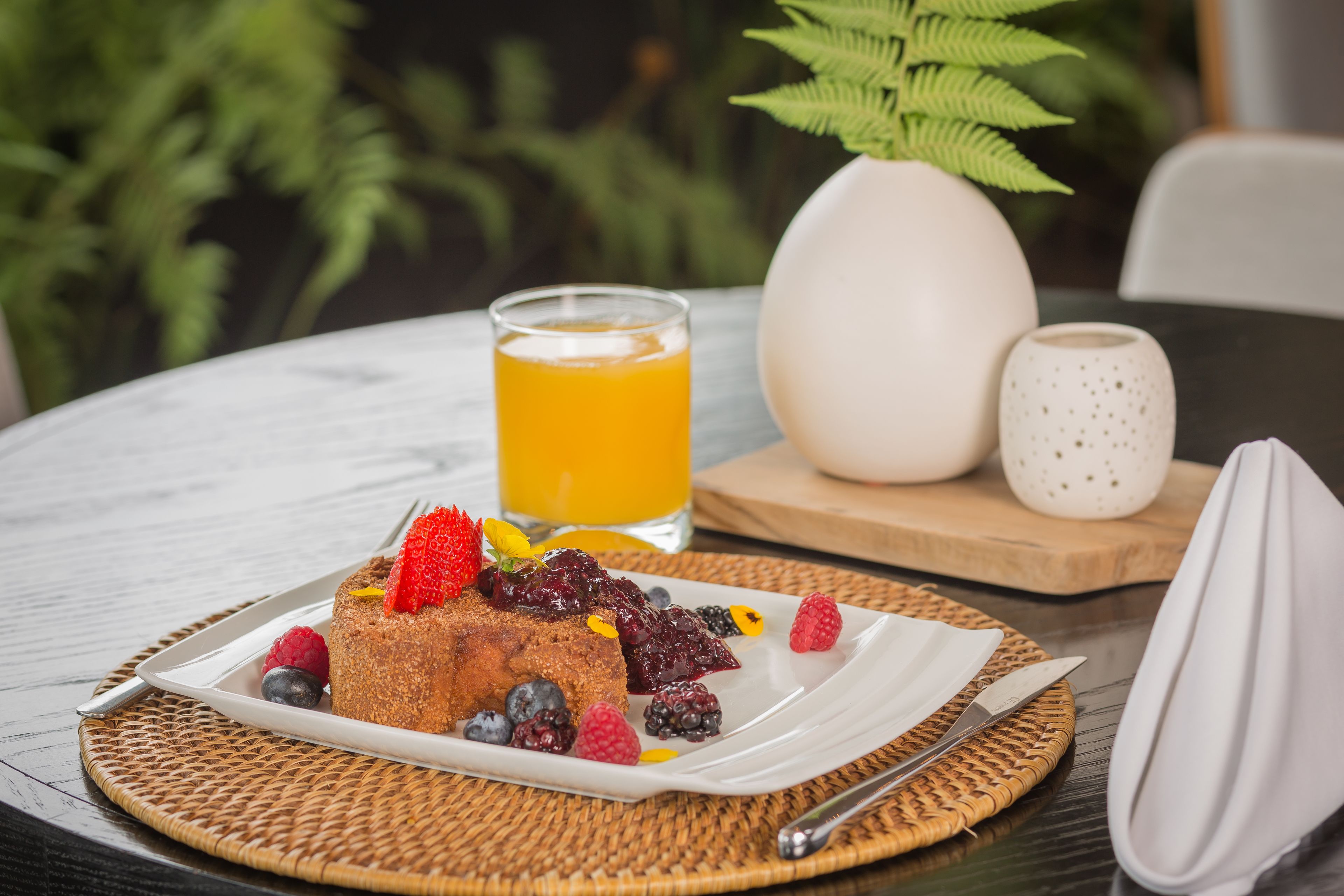 Una escena de desayuno con una fuente de fruta y un vaso de zumo de naranja sobre una mesa.