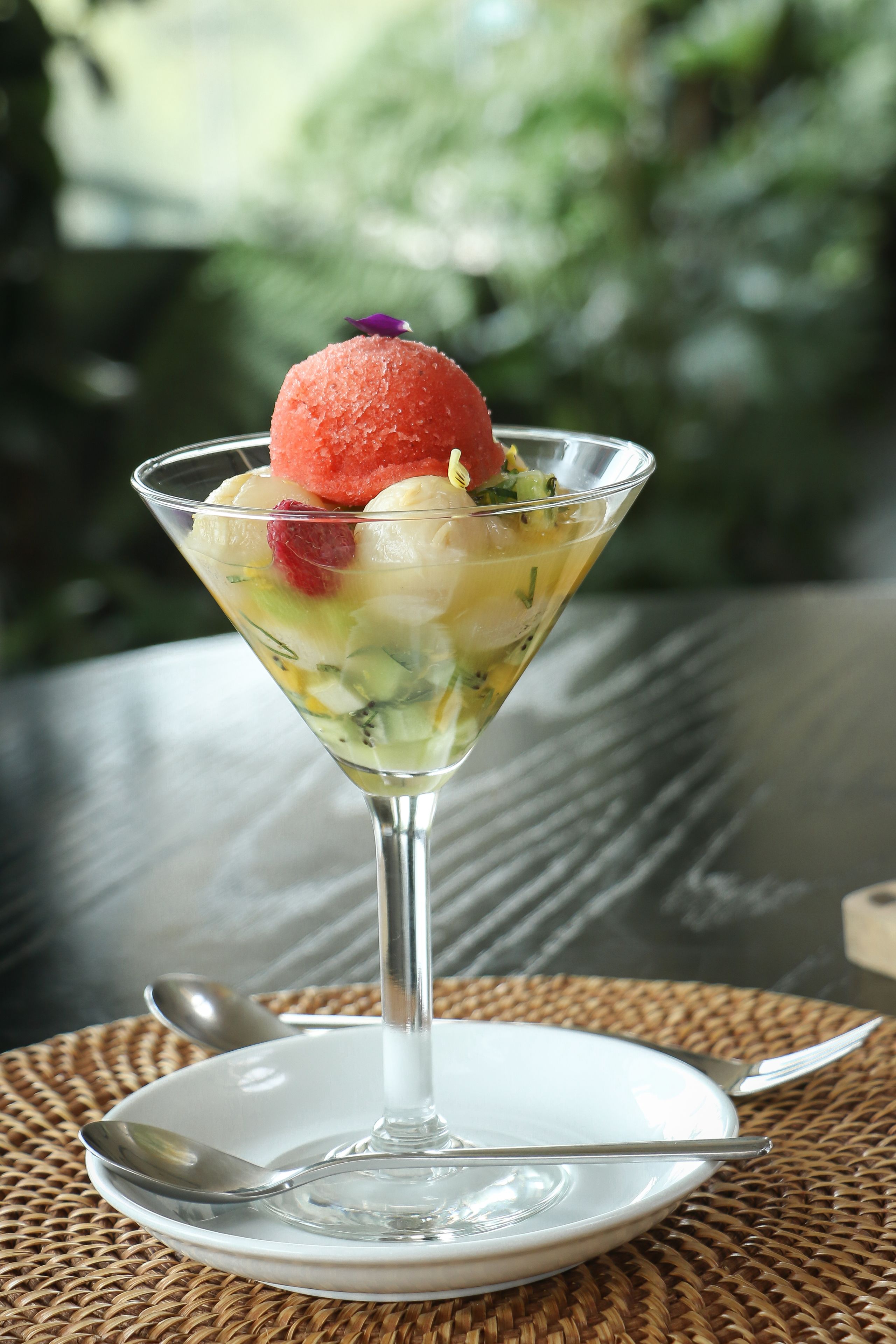 Sobremesa em copo de martini sobre um prato branco, com colher e garfo ao lado, apoiada num guardanapo de tecido.