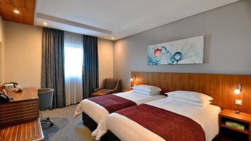 Ein Hotelzimmer mit zwei Betten, einem Schreibtisch, einem Stuhl, einer Mülltonne und einem Bild an der Wand.