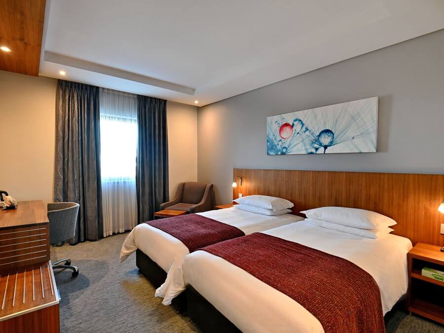 Ein Hotelzimmer mit zwei Betten, einem Schreibtisch, einem Stuhl, einer Mülltonne und einem Bild an der Wand.