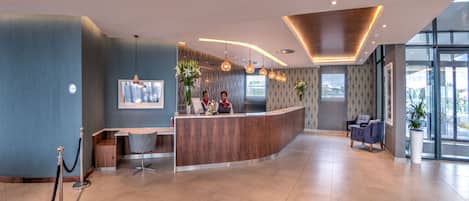 A modern reception area with a curved wooden counter, a seating area with a blue chair, and a large window providing a view of the outdoors.