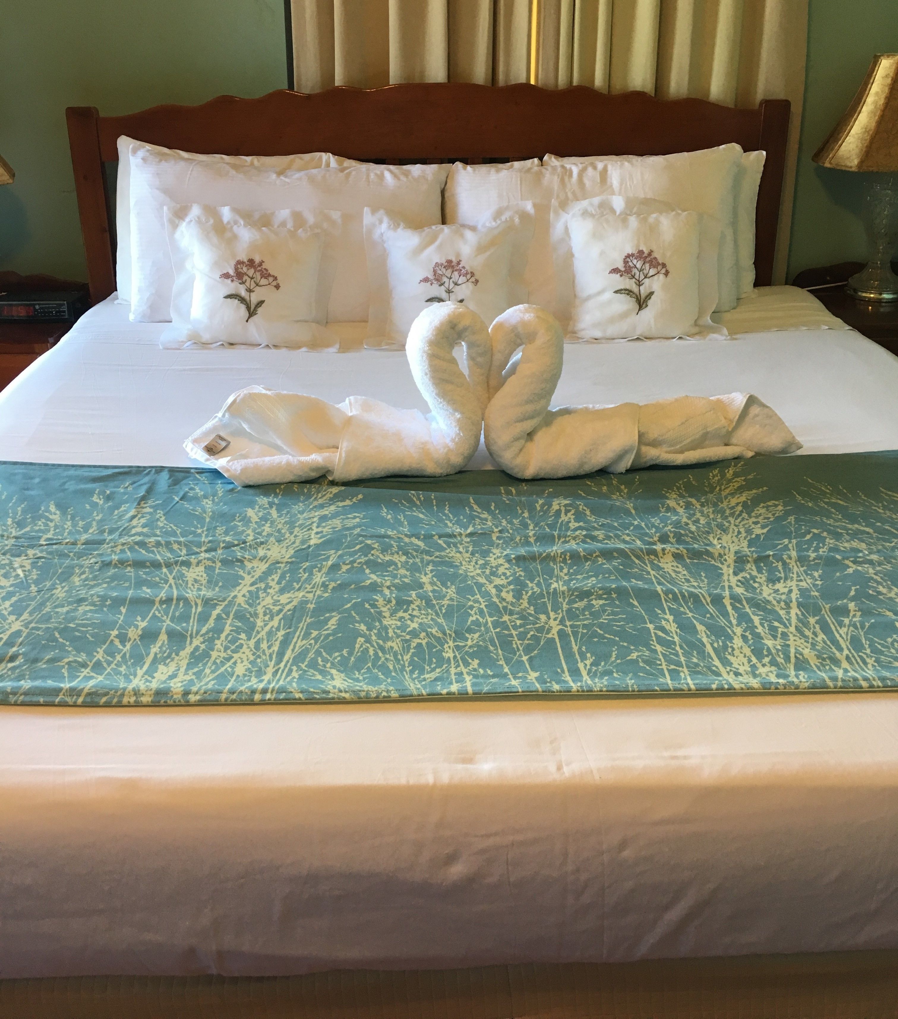 Une chambre d’hôtel avec un lit agrémenté d’une décoration en forme de cygne réalisée avec des serviettes, une couvre-lit à motifs verts et des oreillers blancs ornés de broderies florales.